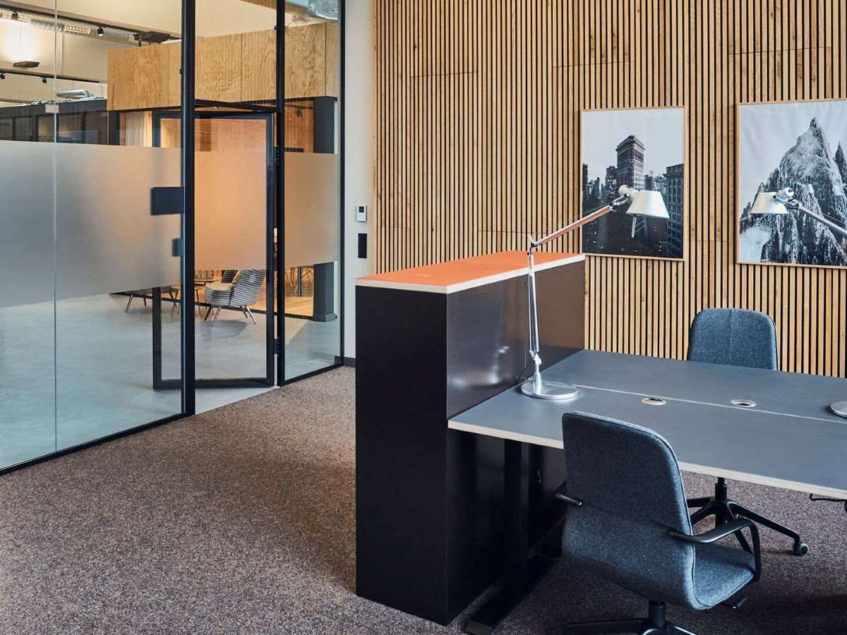Arbeitsplätze in einem Team-Office mit teilweise verglasten, teilweise holzverkleideten Wänden und Teppichboden